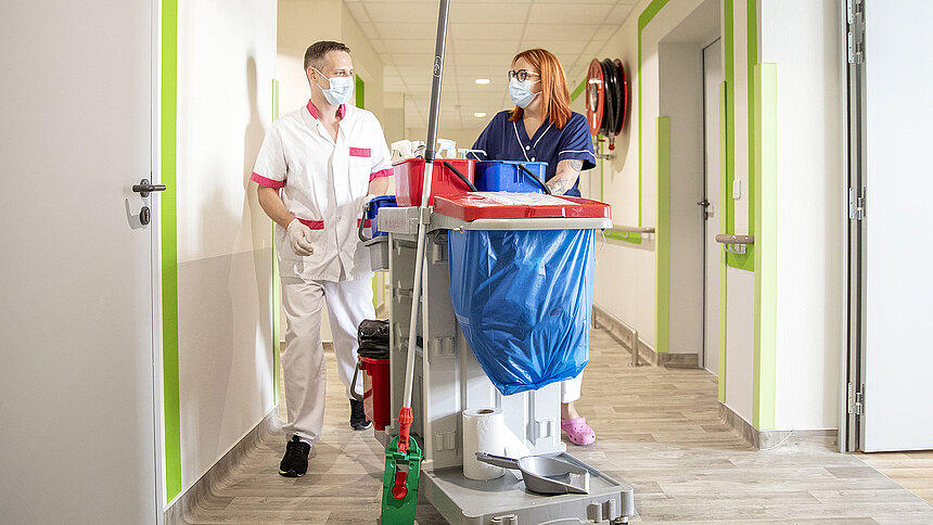 Deux agents de service hospitalier poussent un chariot de ménage dans un couloir.