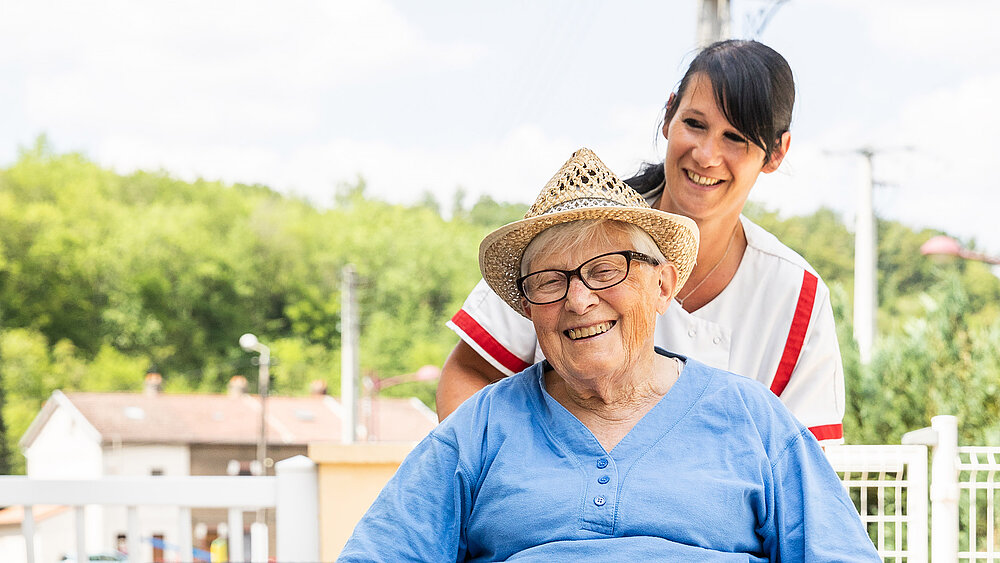 Une aide à domicile promène une personne âgée en fauteuil roulant.