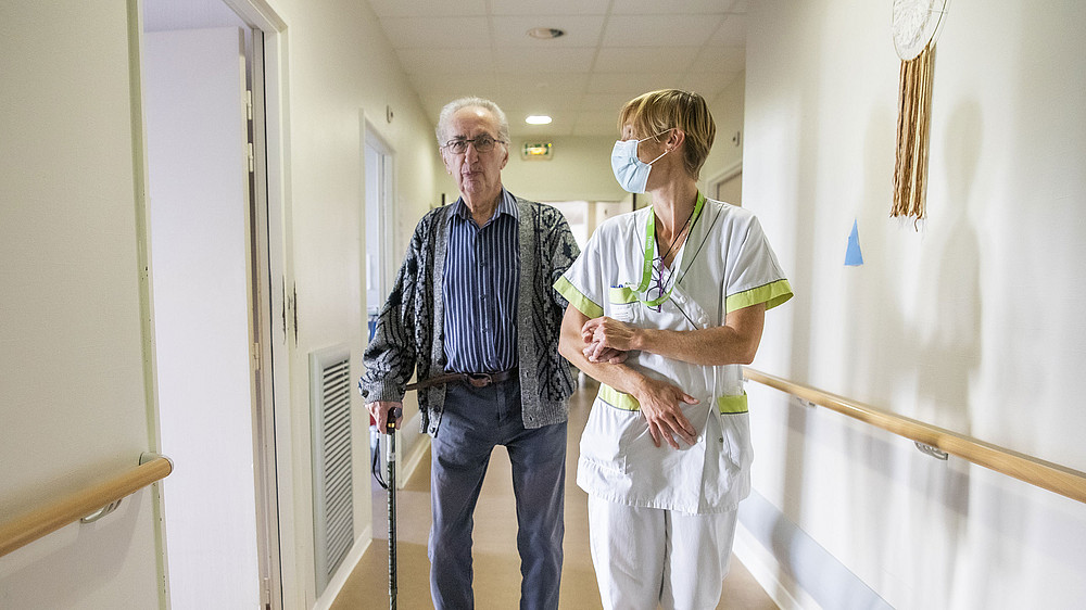 Une professionnelle Filieris tient par le bras un résident dans les couloirs de l'Ehpad Germaine Tillion.