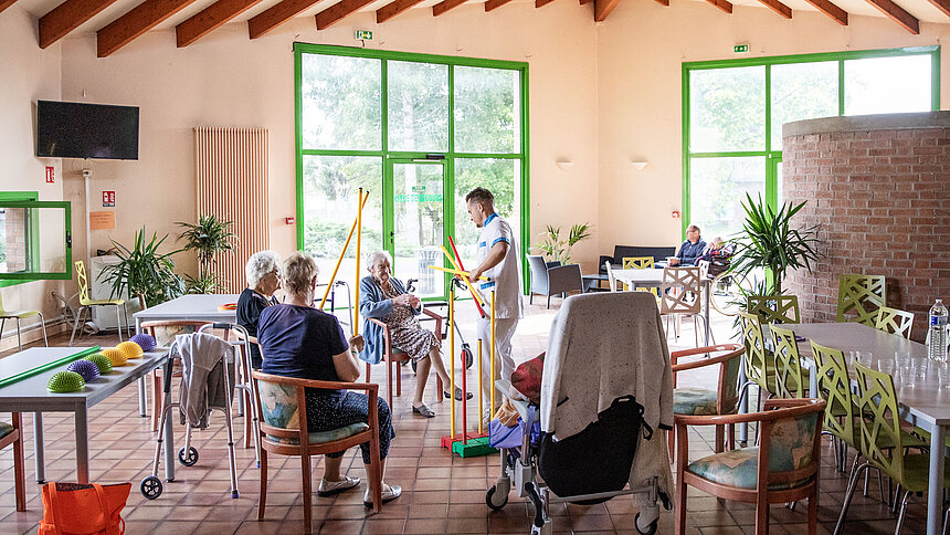 Le kiné guide quatre personnes âgées pour des exercices adaptées dans la salle à manger.